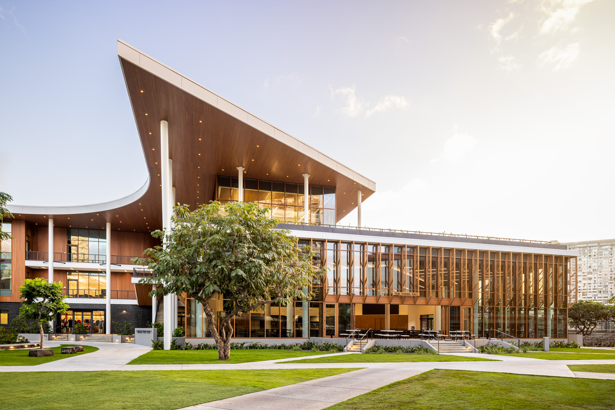 Modern campus building with a sweeping curved roof, warm wood paneling, and large glass windows, surrounded by green lawn and walkways, with a tree in the foreground and soft evening light illuminating the interior.