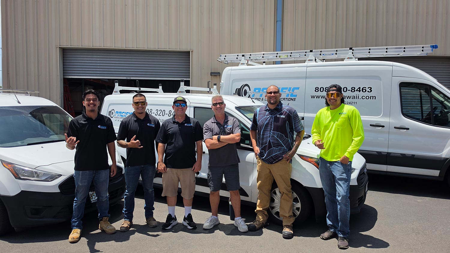 Six men posing in front of two white work vans parked outside a large warehouse building