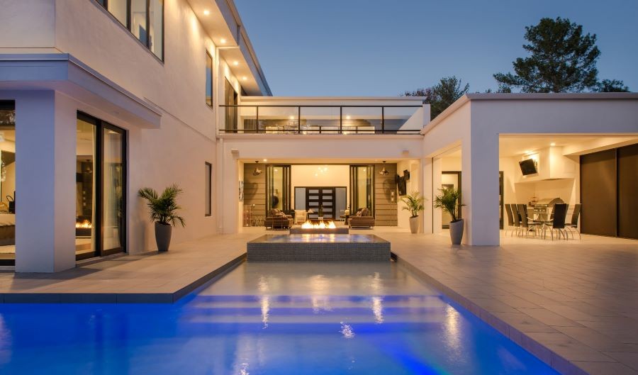 The back view of a well-lit home with a pool and fire feature at dusk.