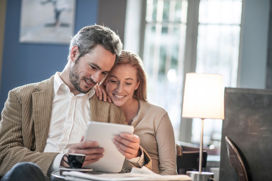 A man in a brown jacket and a woman in a tan cardigan look at an IPad as they sit in their living room.