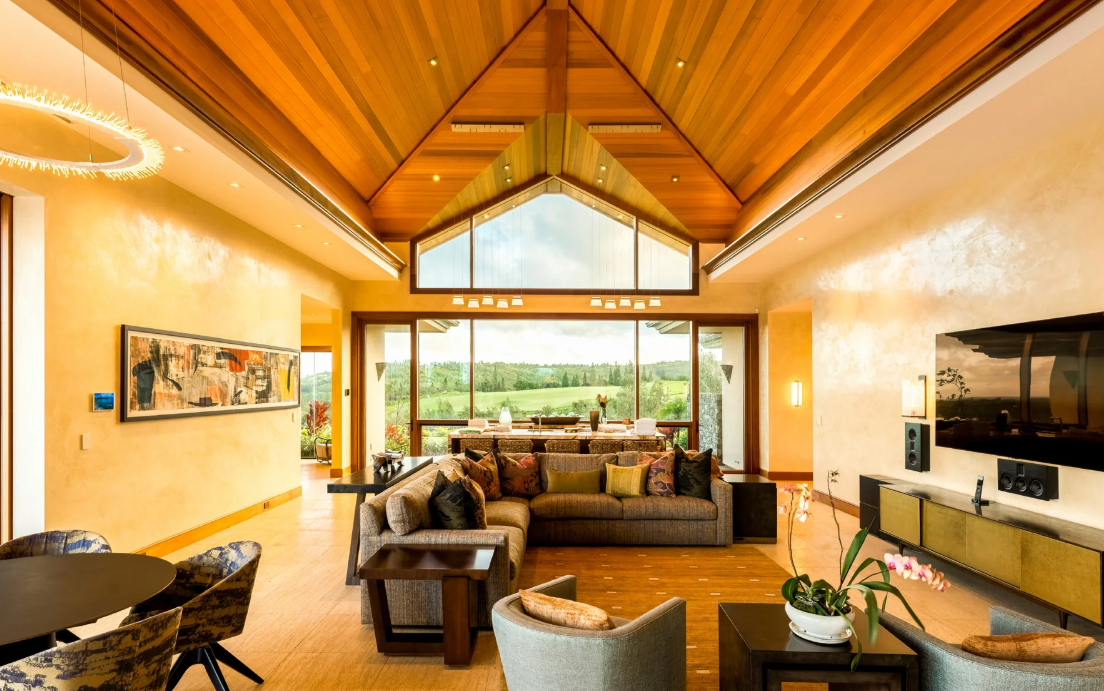 Modern living room with vaulted wood ceiling, large windows overlooking greenery, recessed linear lighting, and neutral-toned furnishings arranged for open-concept living.