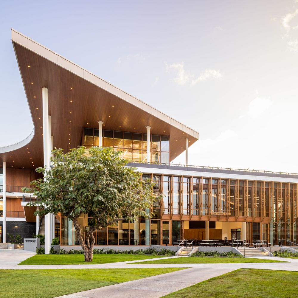 A modern multi-story building with a dramatic, angular roofline and extensive glass walls. The design features warm wood paneling, vertical louvers, and slender white columns. A large tree and manicured lawn with concrete paths sit in the foreground under a clear, bright sky.