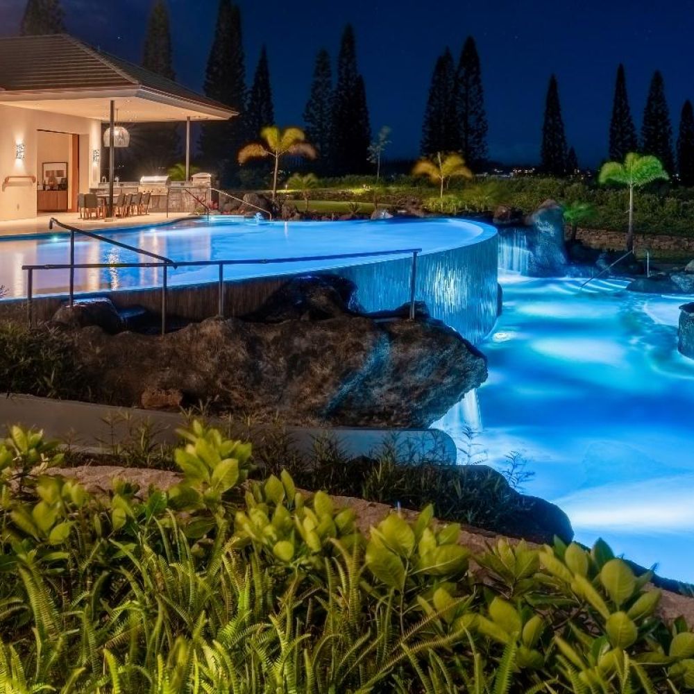 Modern home with illuminated infinity pool and hot tub surrounded by tropical landscaping at night