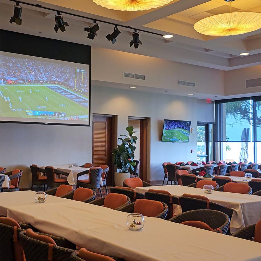 Dining room with long tables, cushioned chairs, and large screens showing a football game
